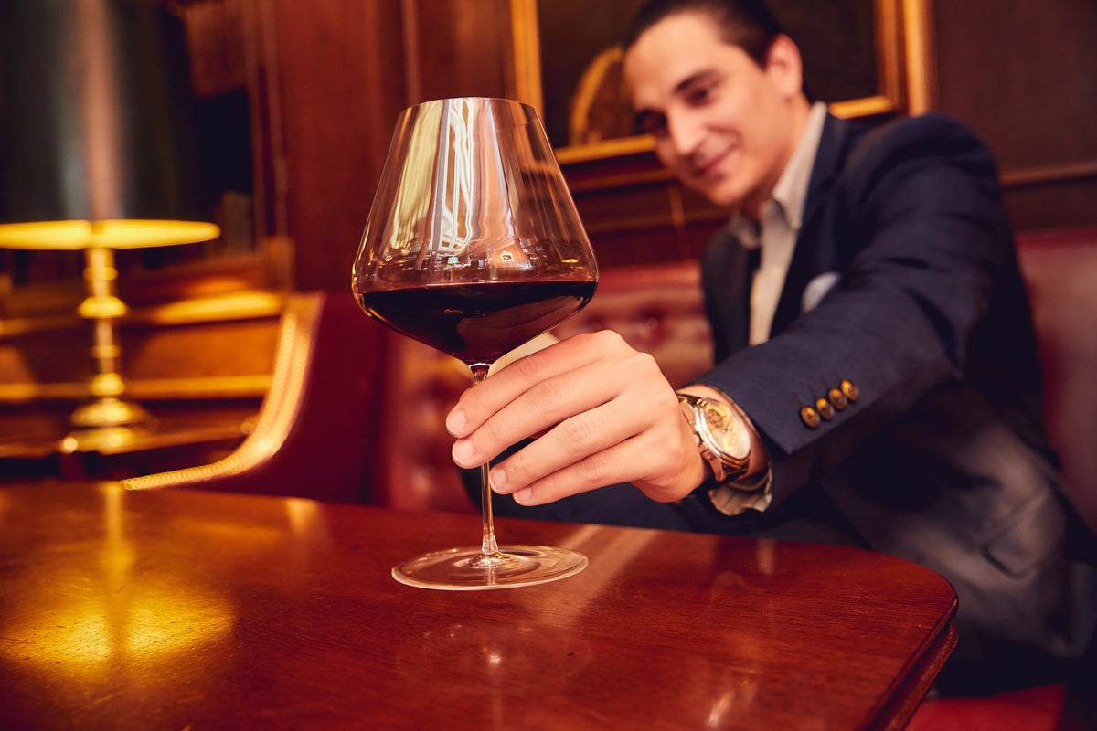 Man presenting a Zalto glass of red wine across a wooden table in a bar